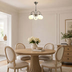 A lit Clérachic chandelier with white glass globes over a round pedestal dining table set with cane-backed chairs and white flowers.