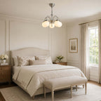 Soft light from this Clérachic chandelier fills a calm cream bedroom with a padded headboard, a linen bench and floral cushions.