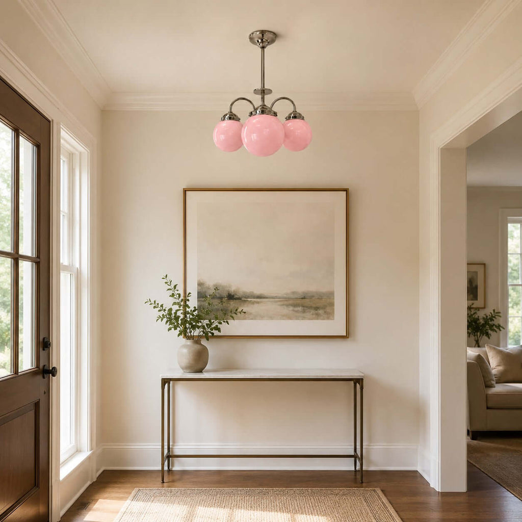 Pink globes on a Clérachic chandelier brighten an entryway with a slim console table, a large framed landscape and a dark wood door.