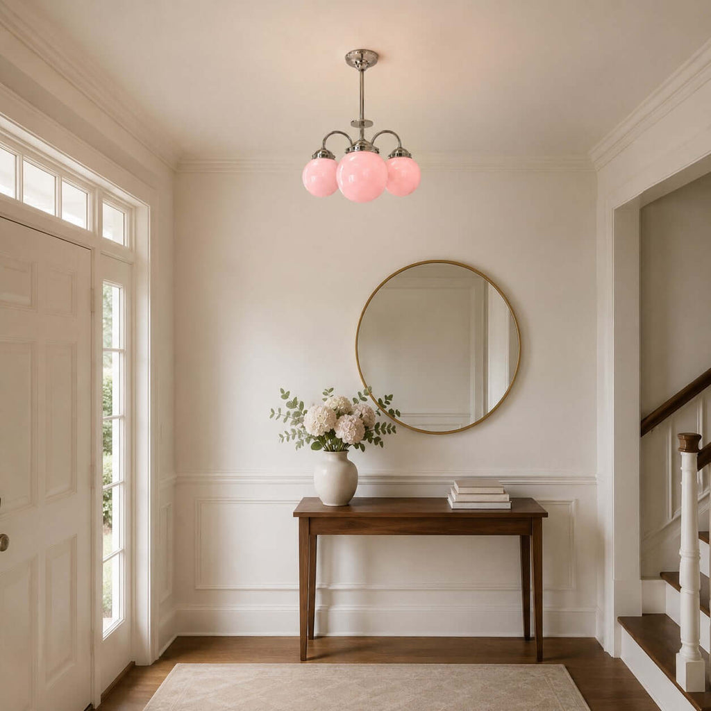 A Clérachic chandelier with three pink globes hangs in a hallway above a wooden console, a round gold-framed mirror and a stair rail.