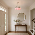 A Clérachic chandelier with three pink globes hangs in a hallway above a wooden console, a round gold-framed mirror and a stair rail.