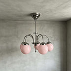Front view of a Clérachic chandelier with pink glass globes, polished metal arms and a stem, hanging against a grey limewash wall.
