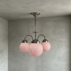 Side view of this Clérachic chandelier with three pink globes on curved polished metal arms, hung from a ceiling plate on a grey limewash wall.