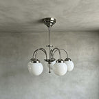Five white glass globes on curved polished metal arms form this Clérachic chandelier, shown hanging from a rod against a grey limewash wall.
