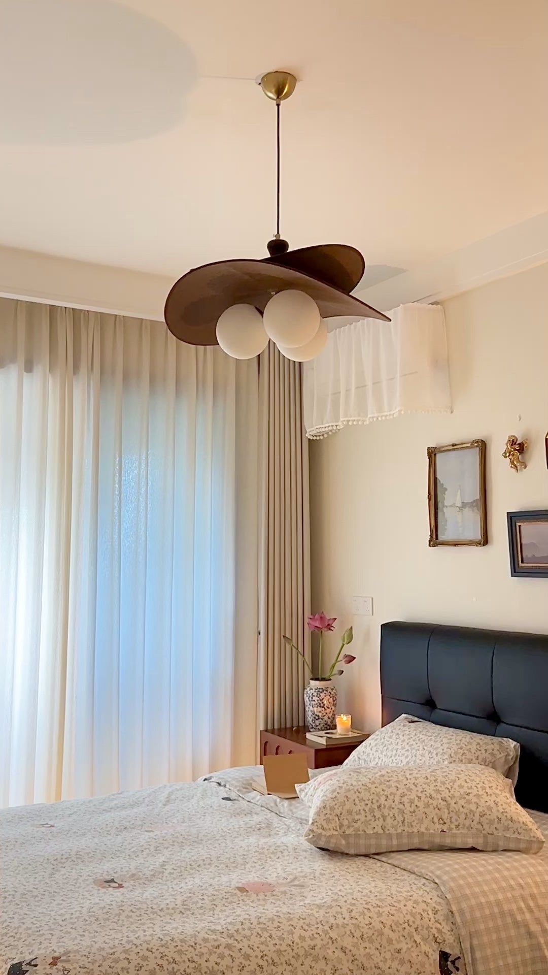 Clérachic pendant with brown petals hung in a bedroom with sheer curtains, a dark headboard and floral pillows below.