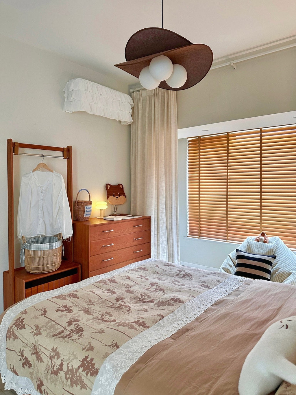 A Clérachic pendant with three globes above a bed with floral bedding, a wooden clothes rail, a chest of drawers and wooden blinds.