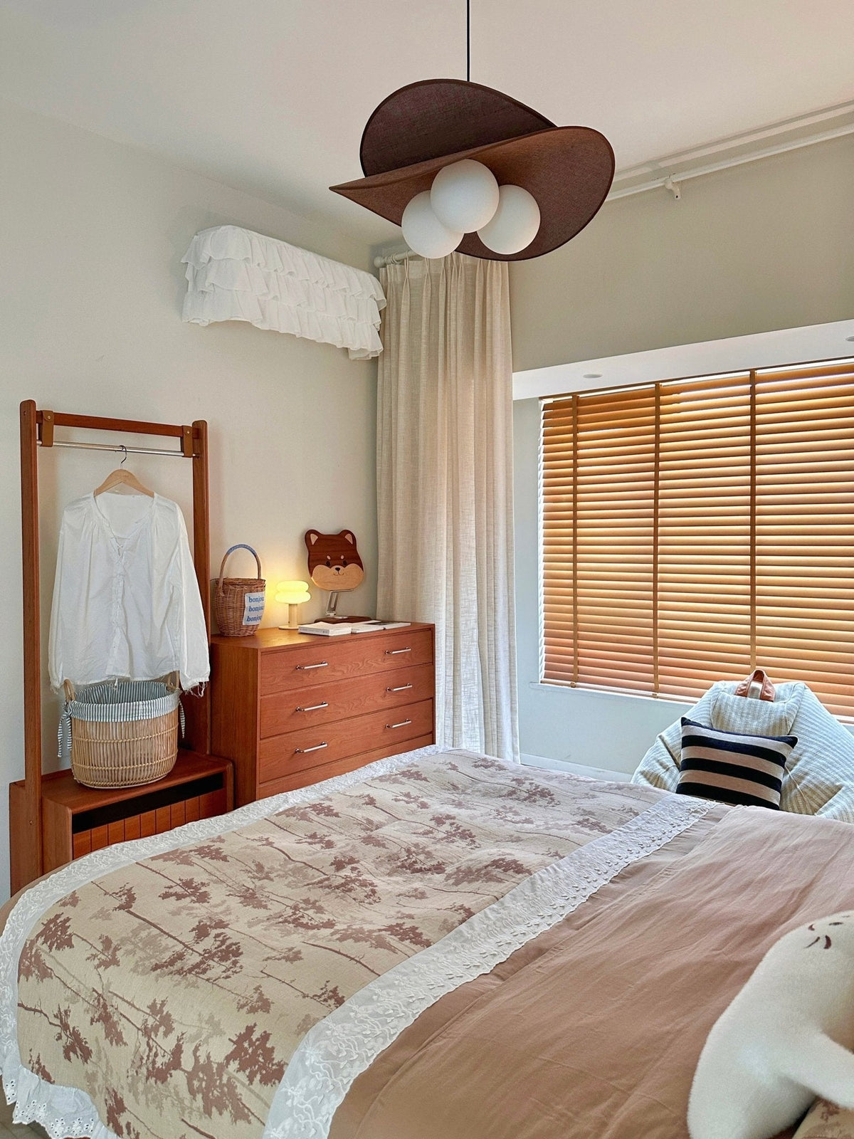 A Clérachic pendant with three globes above a bed with floral bedding, a wooden clothes rail, a chest of drawers and wooden blinds.