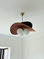 Low angle on this Clérachic pendant, its wide brown fabric shade tilted over three round globes in a plain white room.