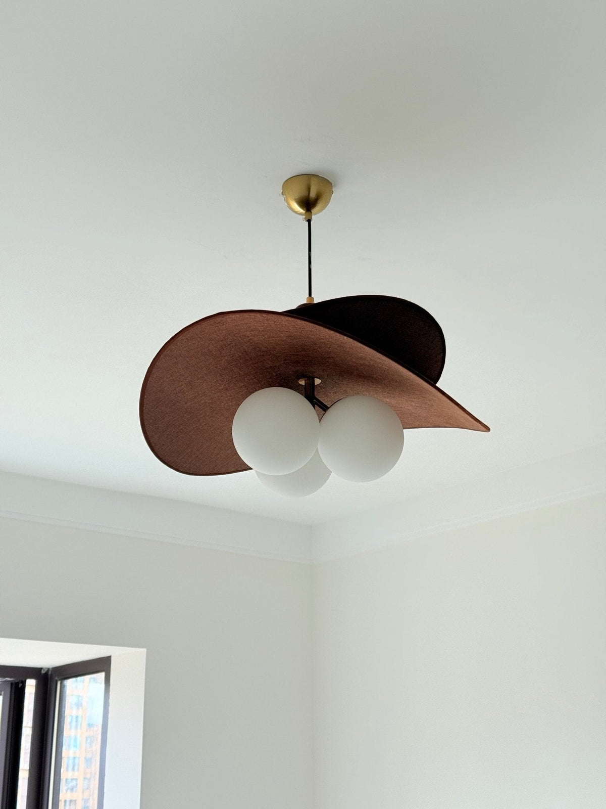 Low angle on this Clérachic pendant, its wide brown fabric shade tilted over three round globes in a plain white room.