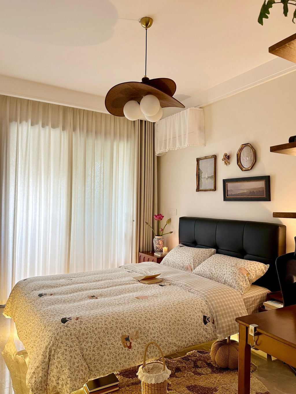 A Clérachic pendant with brown petals hangs beside sheer curtains, above a bed with a black headboard and a patterned quilt.