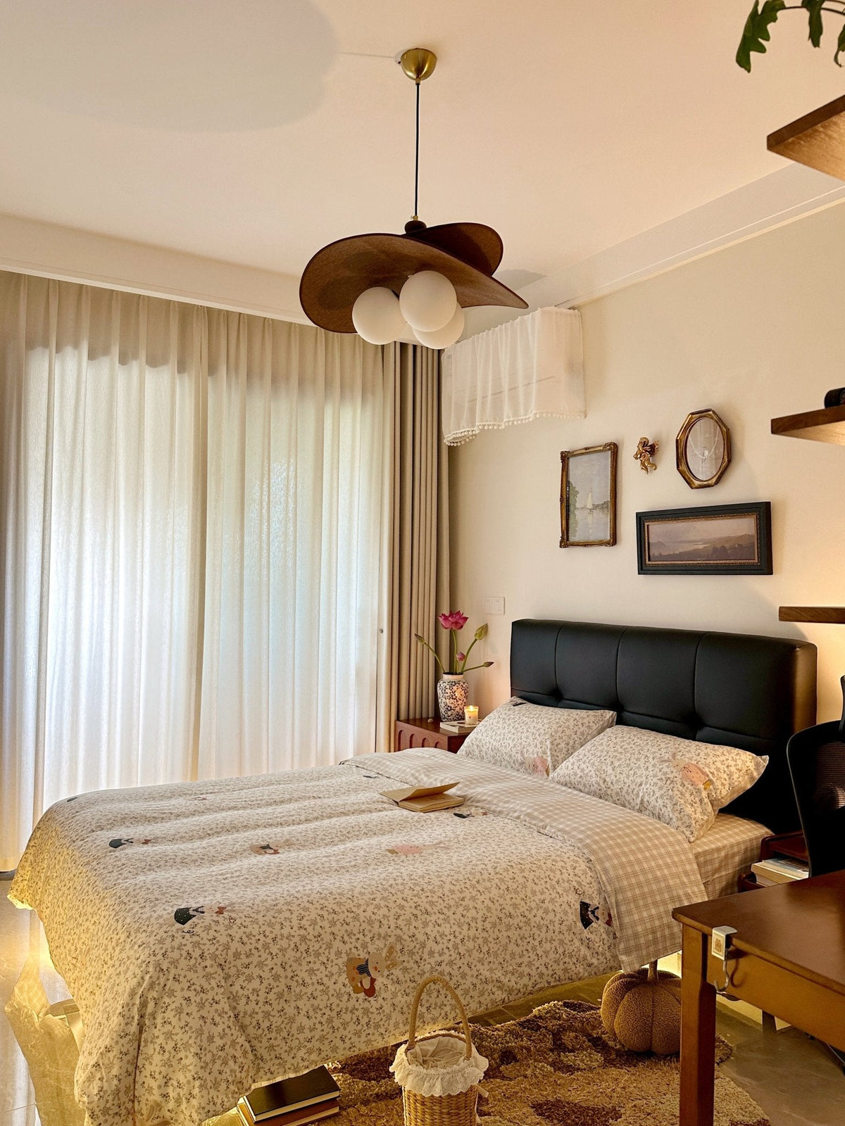 A Clérachic pendant with brown petals hangs beside sheer curtains, above a bed with a black headboard and a patterned quilt.