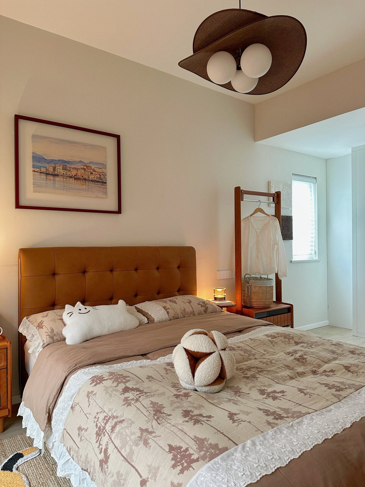 This Clérachic pendant sits at the top of a bedroom with a tan tufted headboard, a framed harbour print and a plush cat toy on the bed.