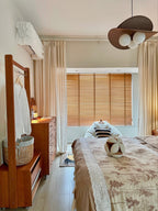A Clérachic pendant at the top right of a bedroom with a clothes rail, a chest of drawers, wooden blinds and a patterned throw.