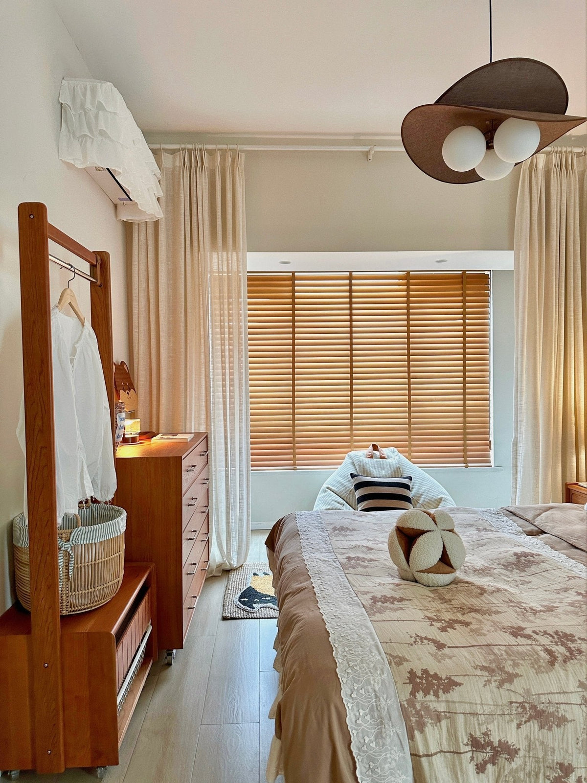 A Clérachic pendant at the top right of a bedroom with a clothes rail, a chest of drawers, wooden blinds and a patterned throw.