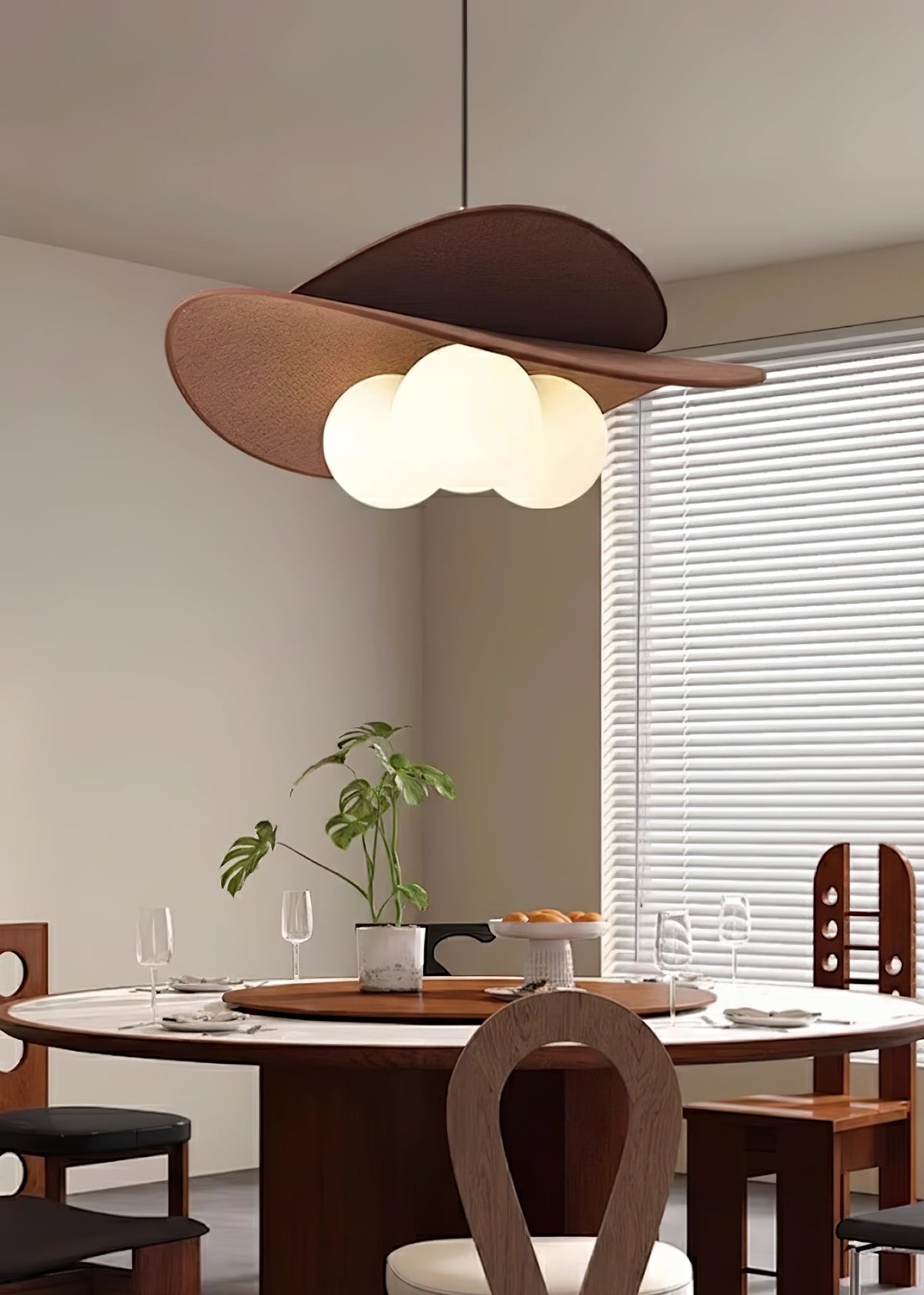 Clérachic pendant with a warm glow above a round dining table with dark wood chairs, a plant and a window with white blinds.