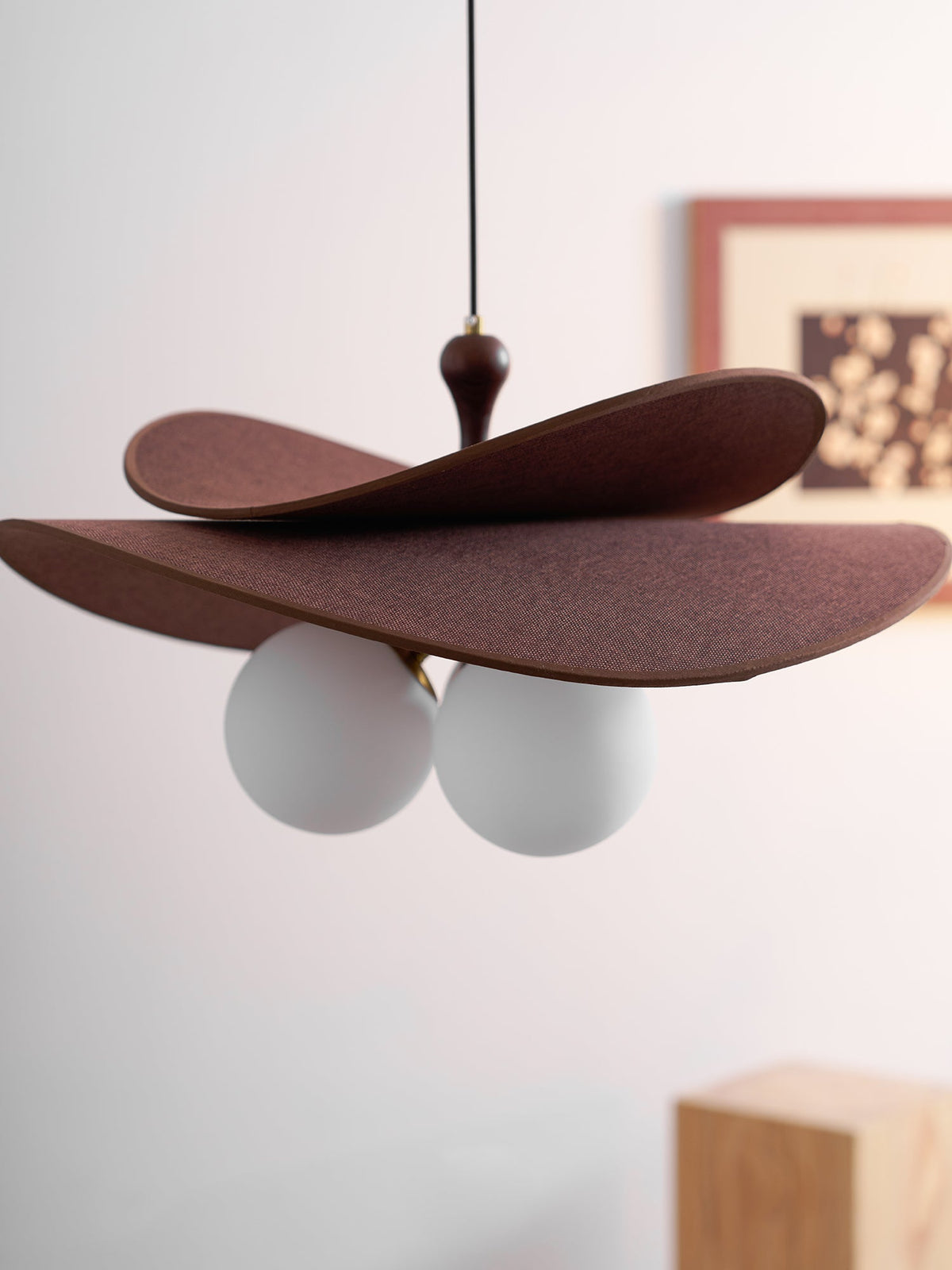 Side view of this Clérachic pendant with two brown petals tilted over white globes, in soft focus against a pale wall and a framed print.