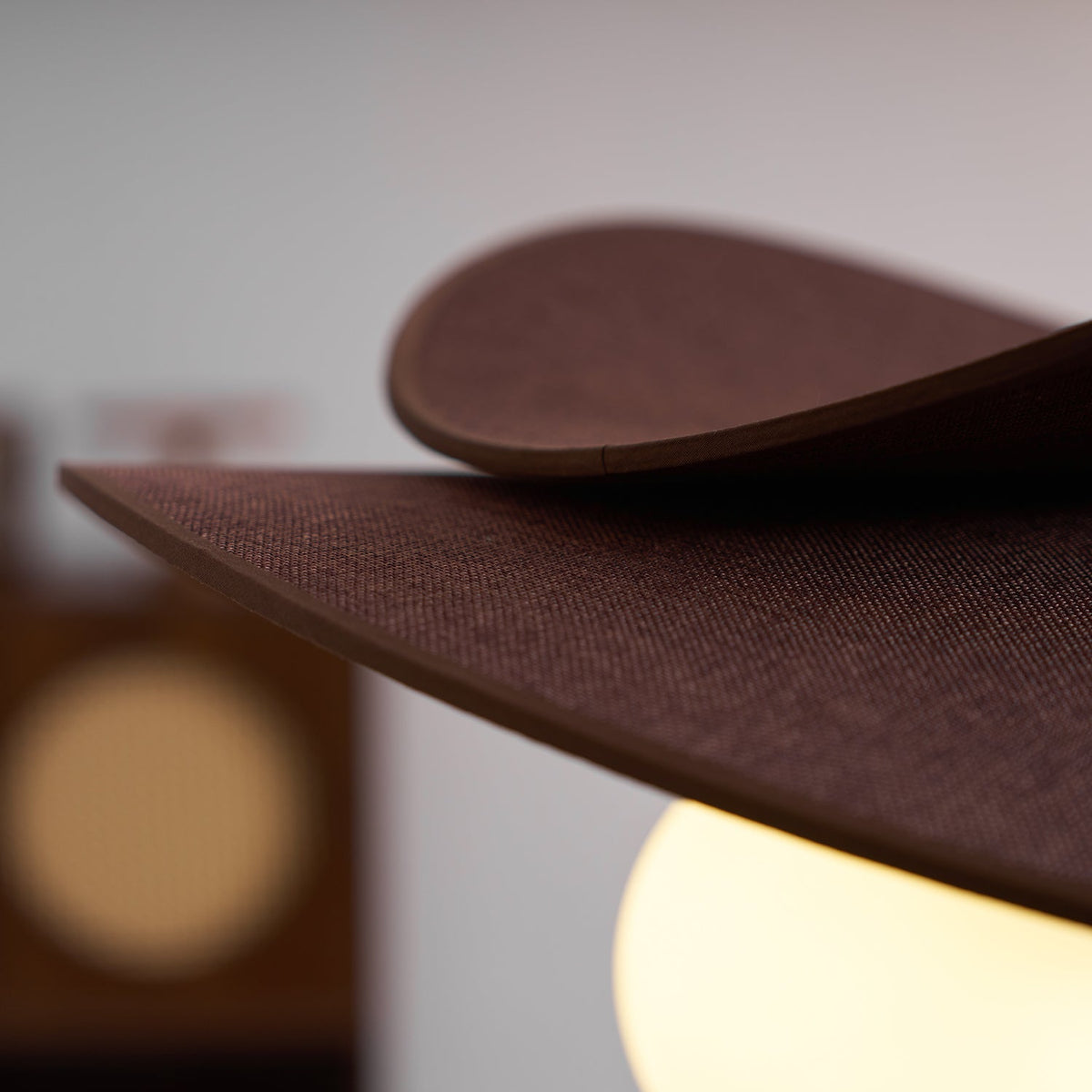 Close view of the layered edge of the brown fabric petals and a lit globe on this Clérachic pendant, with a blurred room behind.