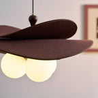 Underside of this Clérachic pendant with three lit globes glowing warm beneath the brown petals, and a framed print blurred behind.