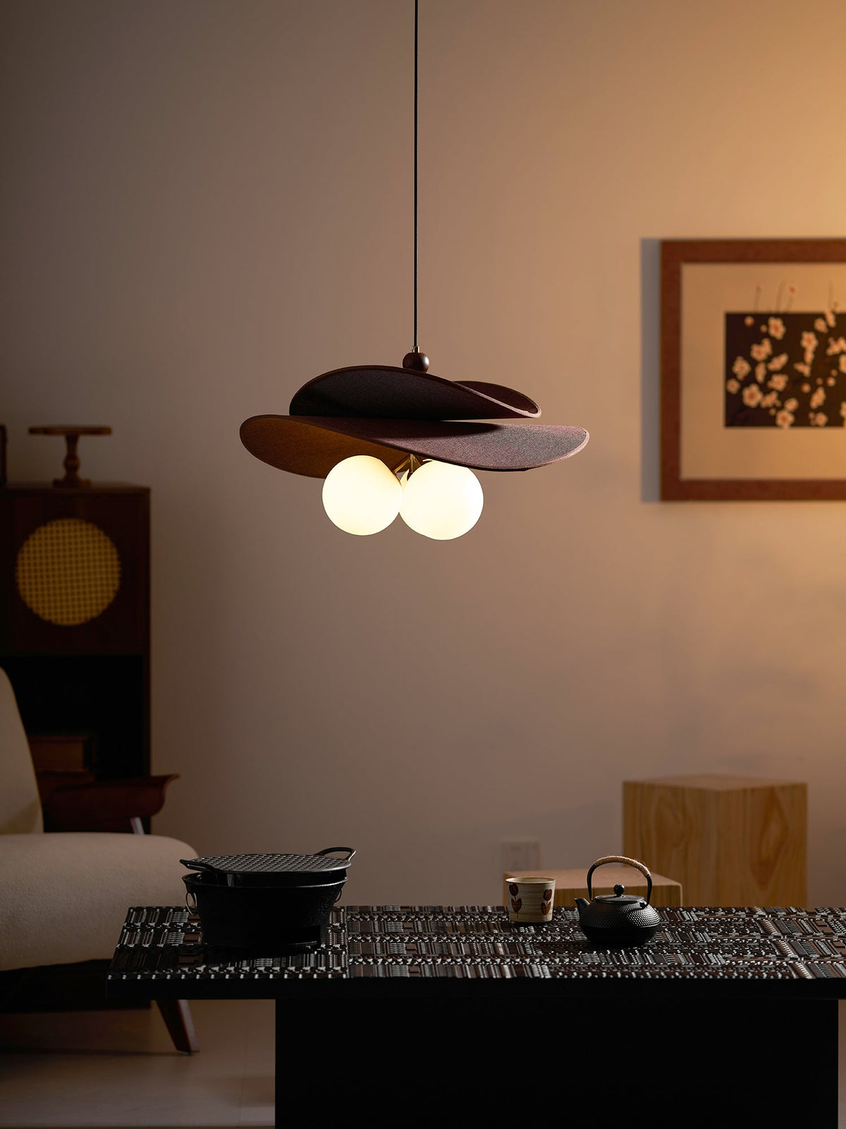 Clérachic pendant glowing in evening light above a dark low table with a teapot, with a framed print on the wall behind.