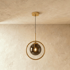 Clérachic gold ring pendant with a smoky ash glass globe and a filament bulb, hung on a thin cord in a studio