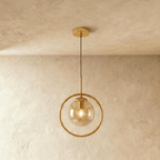 Clérachic gold ring pendant holding an amber glass globe with a filament bulb glowing, shown in a studio