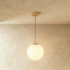 Clérachic gold pendant with a medium white opal glass globe on a slim cord below a round gold canopy, in a studio
