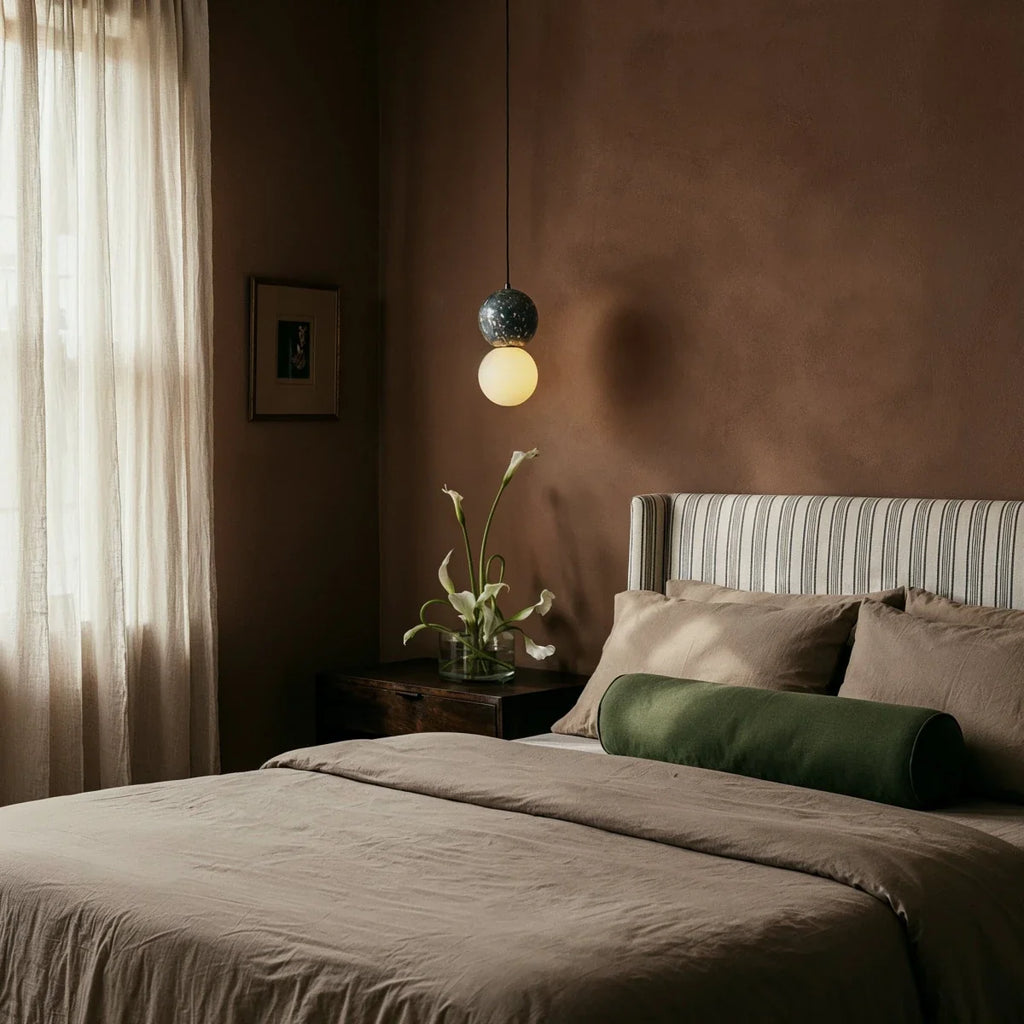 Clérachic two-globe pendant with a dark terrazzo sphere and glowing opal globe hung beside a bed in a warm clay-toned bedroom.