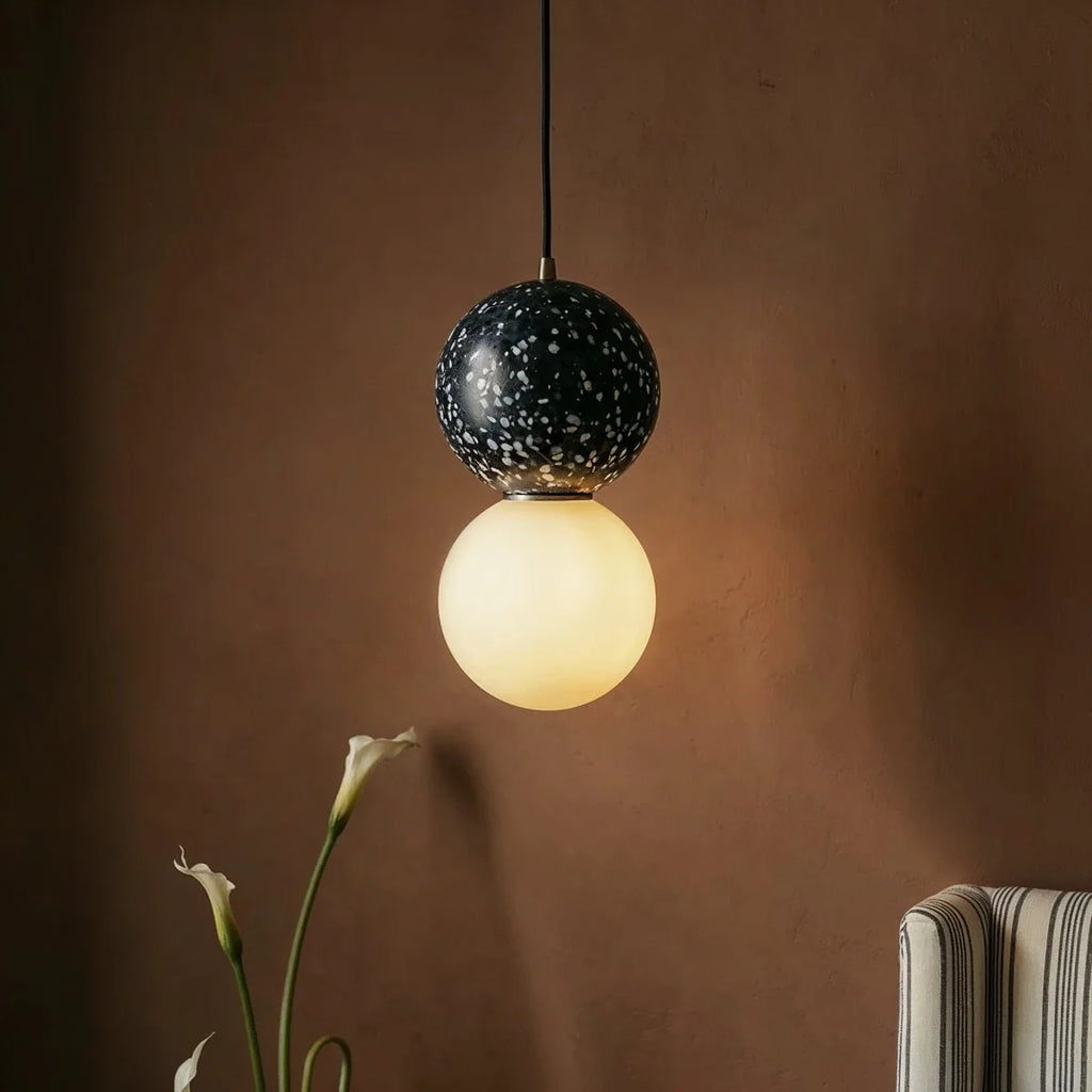 Close view of a Clérachic pendant with a black terrazzo sphere flecked in white above a softly glowing opal glass globe.