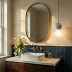Clérachic two-globe pendant with a grey terrazzo sphere hung beside a brass-framed oval mirror above a marble vanity.