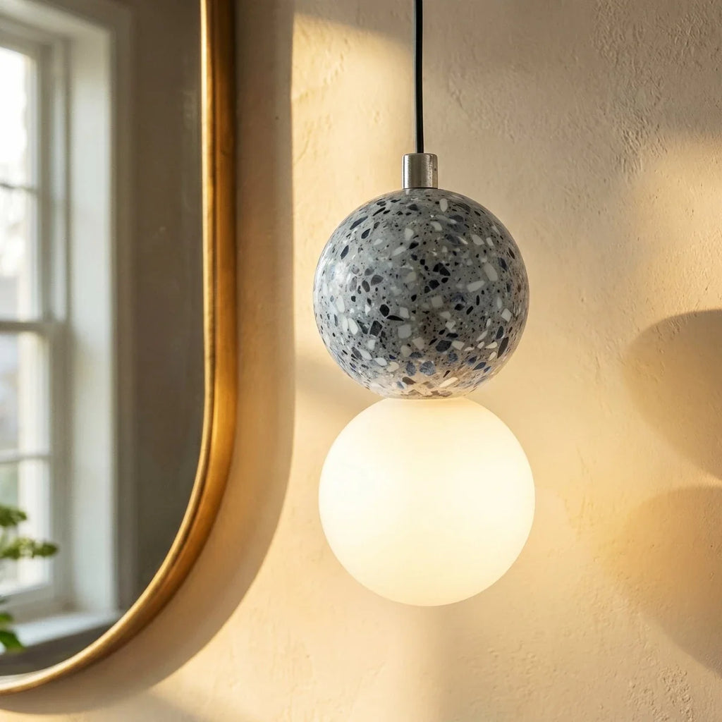 Close view of a Clérachic pendant: grey terrazzo sphere with a chrome cap above a glowing opal glass globe near a brass mirror.