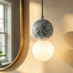 Close view of a Clérachic pendant: grey terrazzo sphere with a chrome cap above a glowing opal glass globe near a brass mirror.