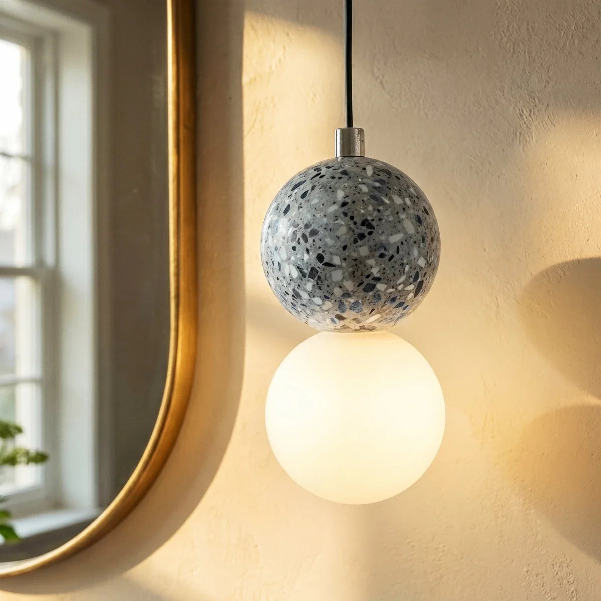 Close view of a Clérachic pendant: grey terrazzo sphere with a chrome cap above a glowing opal glass globe near a brass mirror.
