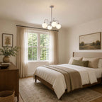 A Clérachic chandelier with chrome arms and white globes above a wooden bed dressed in cream, with sheer curtains at the window.