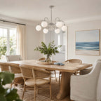 Over a pale oak dining table with woven chairs, a Clérachic chandelier hangs near a window and a blue seascape.