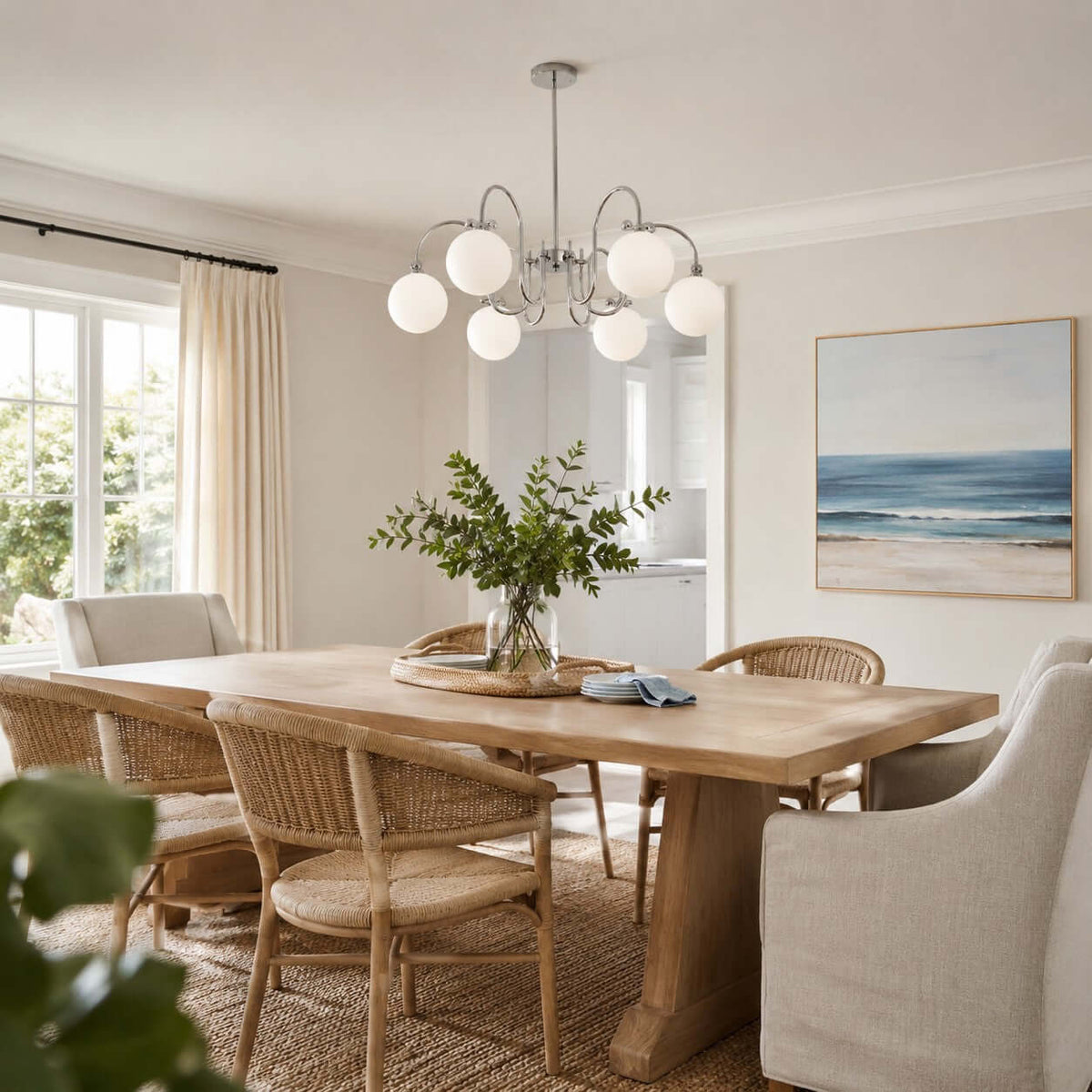 Over a pale oak dining table with woven chairs, a Clérachic chandelier hangs near a window and a blue seascape.