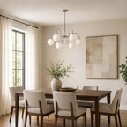 A Clérachic chandelier hangs from a long rod in a dining room with a wooden table, upholstered chairs and a tall window.