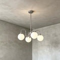 Four frosted globes on curving chrome arms, a Clérachic chandelier hung against a grey plaster wall.