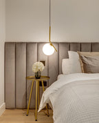 Beside a velvet panelled headboard, a Clérachic pendant with a brass stem hangs over a gold side table holding white flowers.