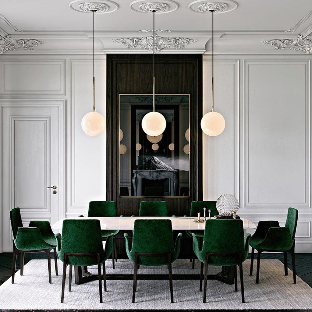 Three Clérachic globe pendants hang in a row over a long dining table with dark-green velvet chairs in a panelled white room.