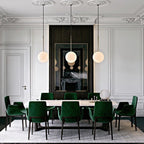 Three Clérachic globe pendants hang in a row over a long dining table with dark-green velvet chairs in a panelled white room.