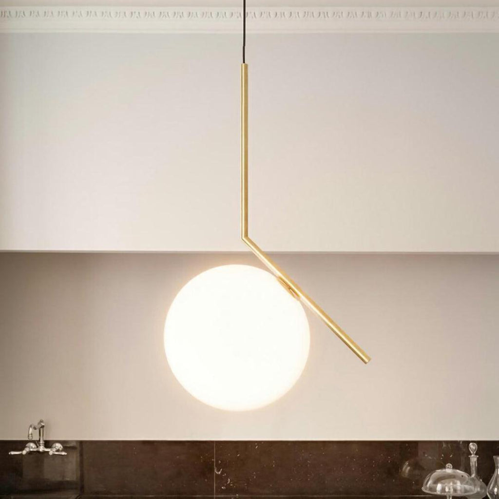 A large Clérachic globe pendant with a brass stem hangs against a cream wall with a plaster cornice above a dark stone counter.
