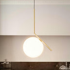 A large Clérachic globe pendant with a brass stem hangs against a cream wall with a plaster cornice above a dark stone counter.