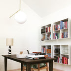 A frosted white Clérachic globe pendant hangs in a white study above a dark wooden desk, with a shelving unit of books behind.