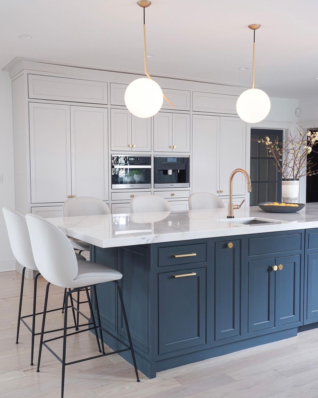 Two Clérachic globe pendants with brass stems hang over a white marble kitchen island with a deep blue base and pale stools.