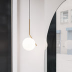 A Clérachic pendant with a brass stem and white glass globe hangs next to a tall arched mirror in a bright, minimal hallway.
