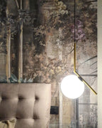A Clérachic globe pendant with a brass stem hangs in front of a floral mural wallpaper and a button-tufted taupe headboard.