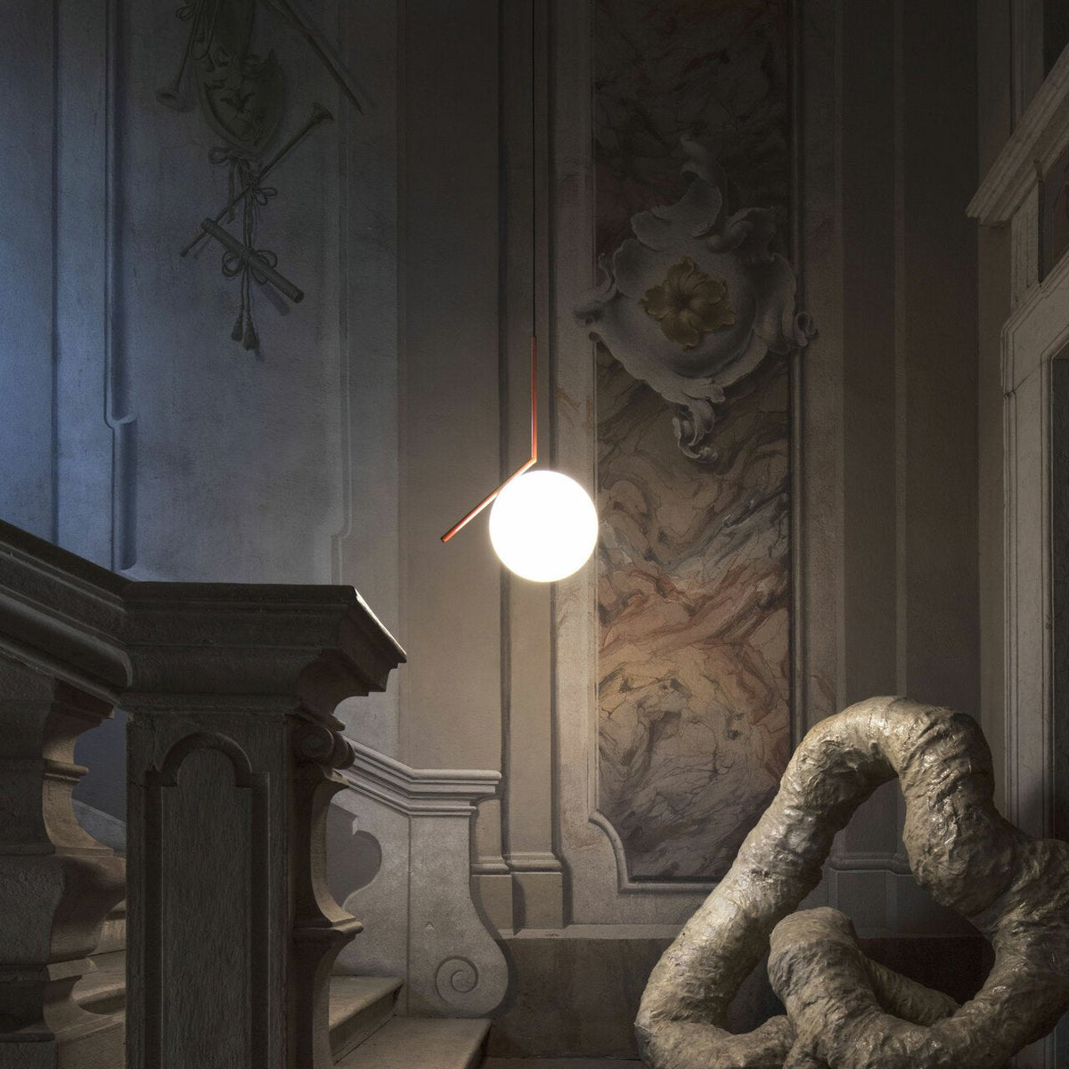 A Clérachic globe pendant glows in a dim stairwell with a stone balustrade, a painted wall and a twisted sculpture below.