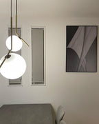 Two Clérachic globe pendants hang in front of a cream wall with narrow windows and a dark abstract print, one globe cropped by the edge.