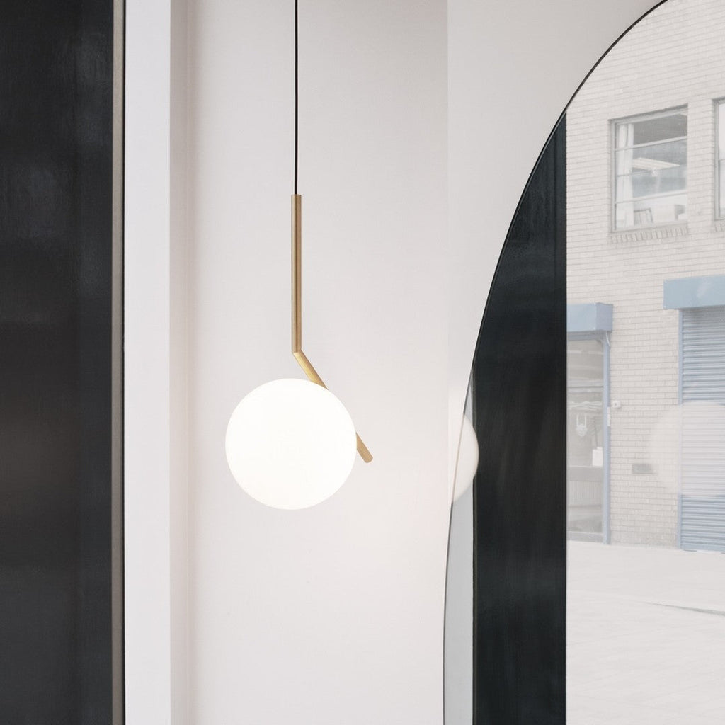 A Clérachic pendant with a brass stem and white globe hangs against a pale wall, with an arched mirror reflecting the street.