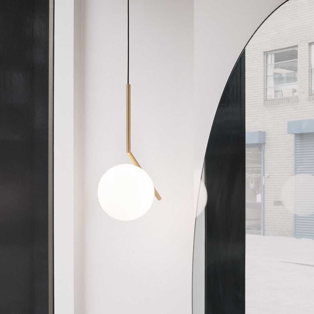A Clérachic pendant with a brass stem and white globe hangs against a pale wall, with an arched mirror reflecting the street.